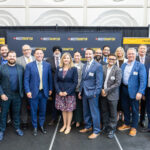 Group of 18 professionals in business attire posing for a photo on a stage with Invest Brampton banners in the background.