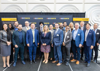 Group of 18 professionals in business attire posing for a photo on a stage with Invest Brampton banners in the background.