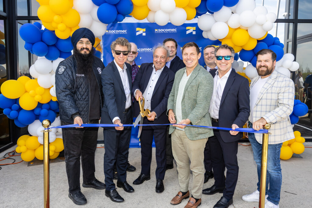 Socomec-36 - Invest Brampton | Brampton Economic Development Group of men in business attire cutting a blue ribbon at a grand-opening event under a colorful balloon arch.