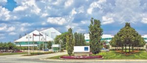 Modern white-roofed office or convention complex with flagpoles, roundabout, and manicured trees under a partly cloudy sky