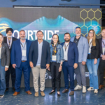 Group of diverse professionals posing for a photo at a conference, in front of a large digital screen backdrop with hexagon graphics.