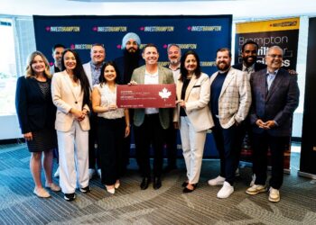 Group of twelve diverse professionals posing in a bright office, holding a large red ceremonial cheque while front banners read 'Invest Brampton'.