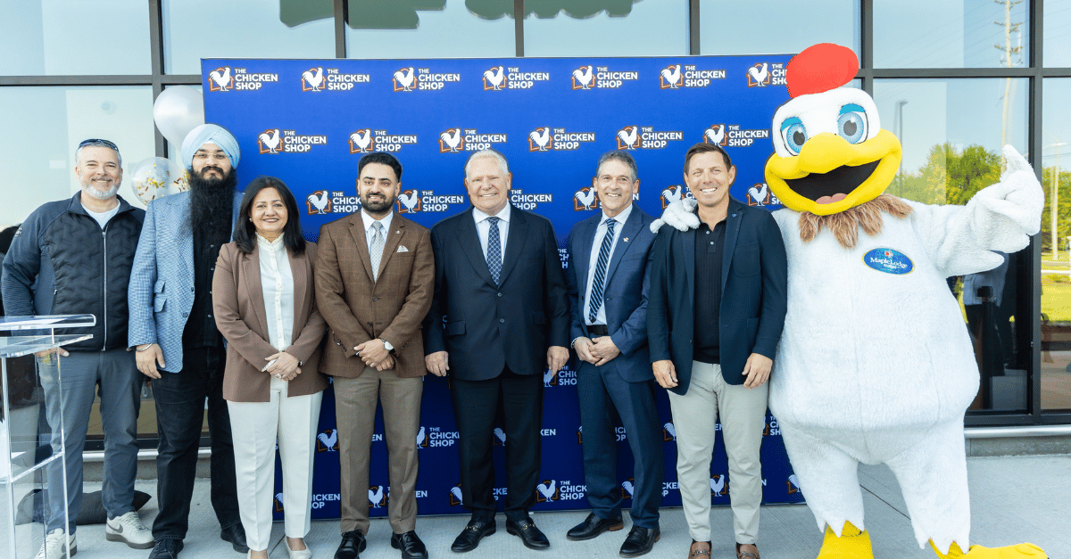Group of seven adults in business attire posing with a chicken mascot in front of a branded backdrop at a The Chicken Shop event.