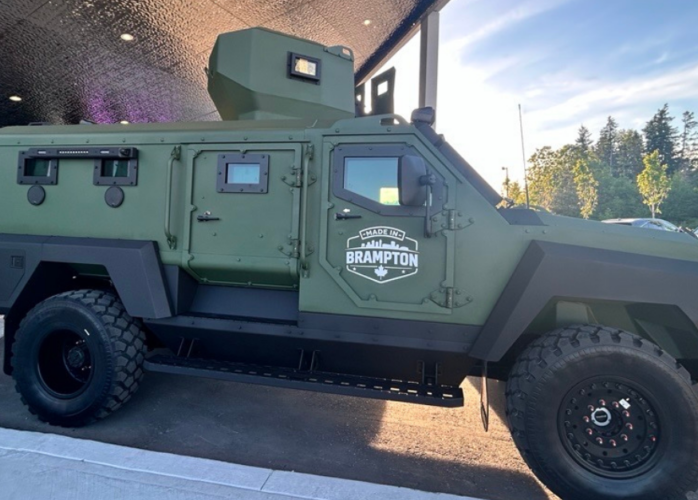 Green armored military-style vehicle with rugged tires parked under a canopy; Brampton emblem on the door.