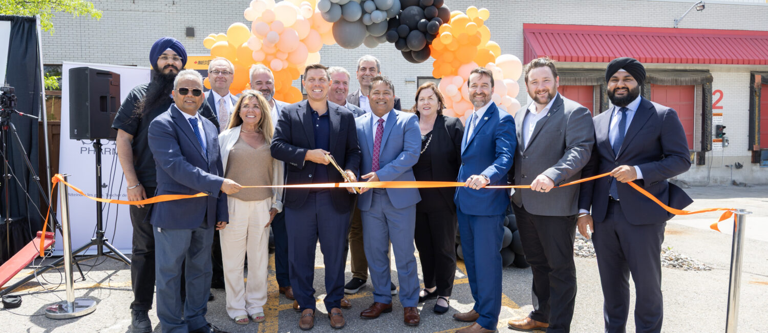 Group of professionals in suits cutting an orange ribbon at an outdoor ceremony with a balloon arch in peach, gray, and black behind them.