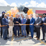 Group of professionals in suits cutting an orange ribbon at an outdoor ceremony with a balloon arch in peach, gray, and black behind them.