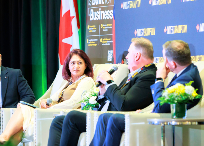 Four panelists seated in cream chairs on a stage, microphones in hand, with a blue Invest Brampton backdrop and a Canadian flag nearby.