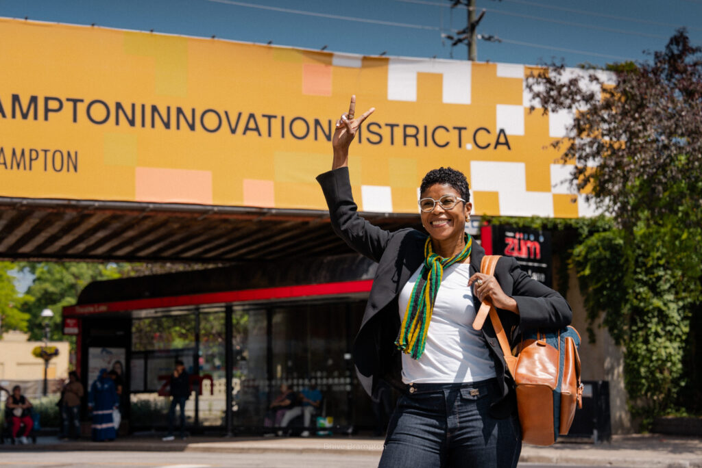 Smiling woman in a blazer with a green scarf raises a peace sign in front of a yellow wall that reads Hampton Innovation District CA.