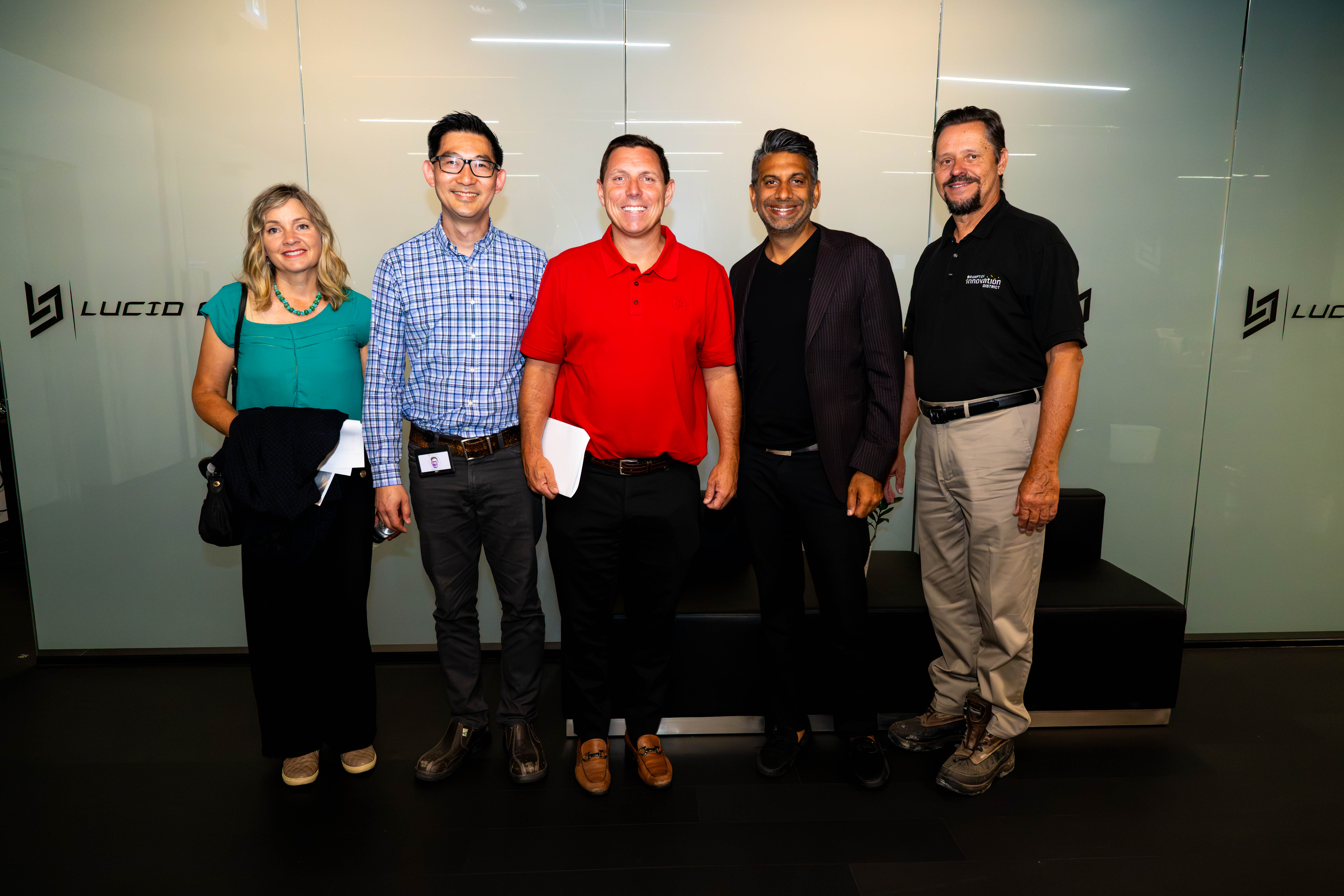 Five professionals pose together in a modern office lobby with glass walls and logos in the background, smiling at the camera.