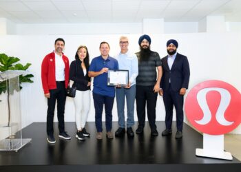 Six adults stand together on a black platform; one man holds a framed certificate as the group smiles, with a large red logo on the right.