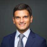 Professional headshot: smiling man in a navy suit and silver tie, dark studio background, corporate use