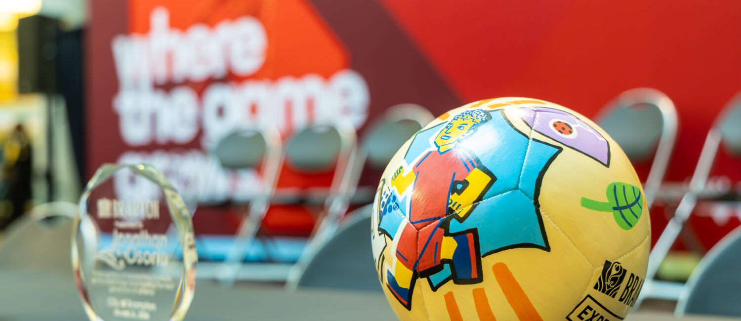 Colorful patterned soccer ball on a stand in a trophy display, with a blurred red backdrop in the background.