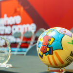 Colorful patterned soccer ball on a stand in a trophy display, with a blurred red backdrop in the background.