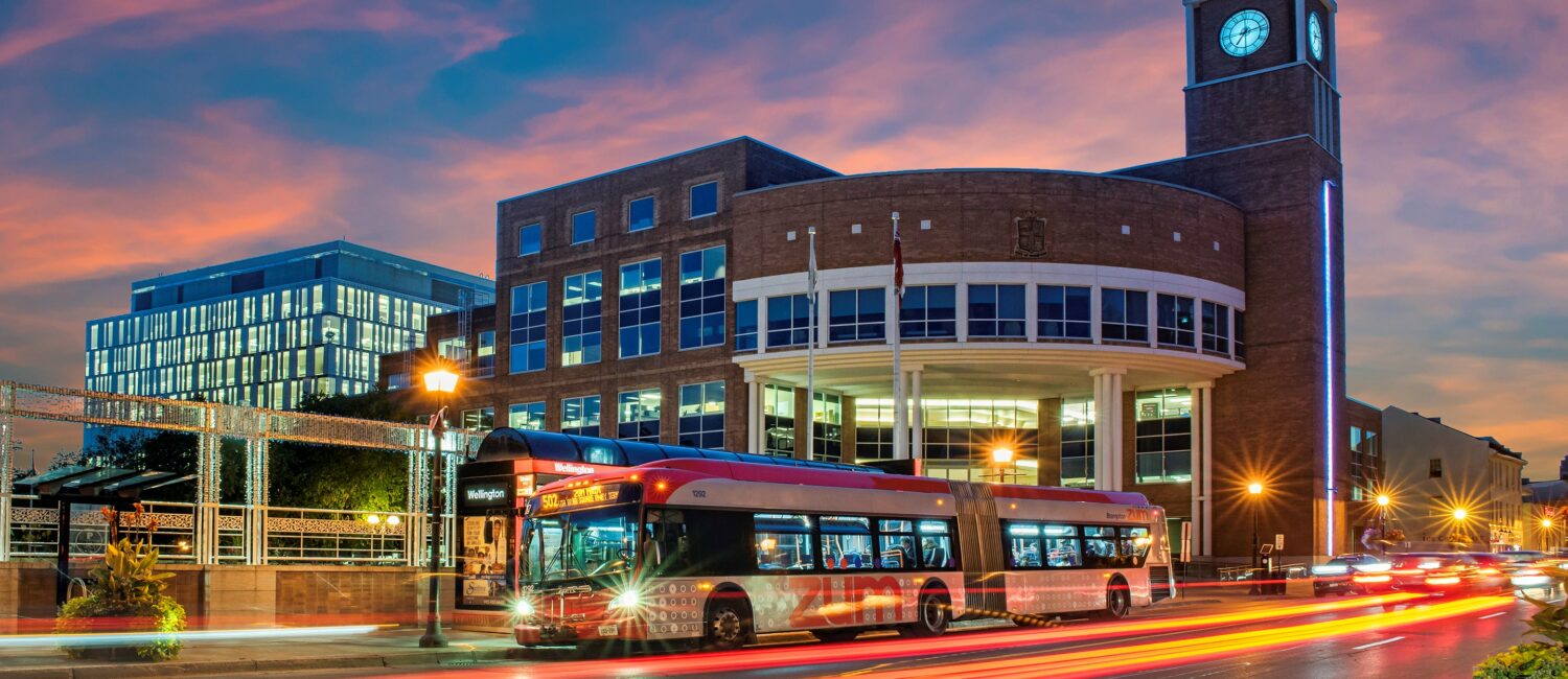 Brick clock-tower building beside a glass-fronted office complex at dusk; a red Wellington bus and light trails on the street.