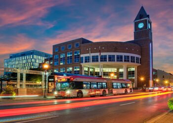 Brick clock-tower building beside a glass-fronted office complex at dusk; a red Wellington bus and light trails on the street.