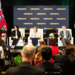 Panel discussion on stage with five speakers, Brampton business banners, and flags, addressing an audience.