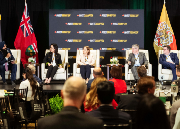 Panel discussion on stage with five speakers, Brampton business banners, and flags, addressing an audience.