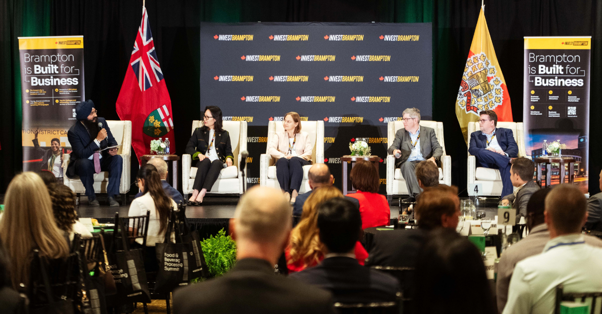 Panel discussion on stage with five speakers, Brampton business banners, and flags, addressing an audience.