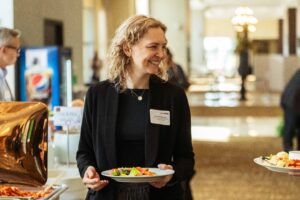 Brampton_Food and Bev Summit_D85_8136 - Invest Brampton | Brampton Economic Development Smiling woman in a black blazer holding a plate of salad at a buffet line in a restaurant. (Name badge visible)