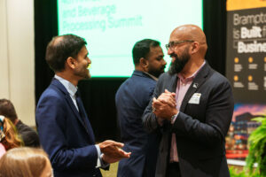 Brampton_Food and Bev Summit_D85_8185 - Invest Brampton | Brampton Economic Development Two men in suits shake hands and smile at a conference, with a bright green presentation screen behind them.