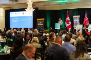 Brampton_Food and Bev Summit_D85_8363 - Invest Brampton | Brampton Economic Development Speaker at a podium on a stage during a formal conference, with a large blue presentation screen, banners, and flags (Canada and UK) in the background, and attendees seated at tables in a banquet-style hall.