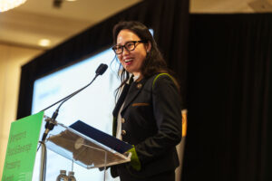 Brampton_Food and Bev Summit_D85_8401 - Invest Brampton | Brampton Economic Development Smiling woman in a dark blazer at a podium, speaking into a microphone with green signage nearby.