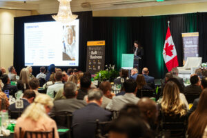Brampton_Food and Bev Summit_D85_8413 - Invest Brampton | Brampton Economic Development Conference hall with a speaker at a podium presenting to an audience; large projection screen and Brampton banners on stage, Canadian flag nearby