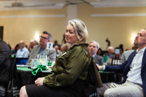 Brampton_Food and Bev Summit_D85_8440 - Invest Brampton | Brampton Economic Development Smiling woman with blonde hair in an olive-green jacket seated at a table at a conference or gala, with other attendees in the background.