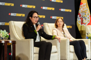 Brampton_Food and Bev Summit_D85_8661 - Invest Brampton | Brampton Economic Development Two women sit on a stage in panel chairs; one speaks into a microphone while the other listens, with an Invest Brampton backdrop behind them.