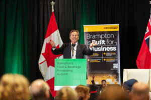 Brampton_Food and Bev Summit_D85_8819 - Invest Brampton | Brampton Economic Development Speaker in a dark suit at a podium on a stage, Brampton banner reading'Brampton's Food & Beverage Processing Summit' visible, Canadian flag behind left side.
