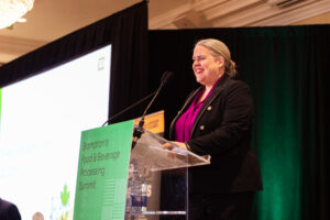 Brampton_Food and Bev Summit_D85_8963 - Invest Brampton | Brampton Economic Development Smiling woman in a purple blouse and black blazer speaks at a conference podium with green signage in the foreground.