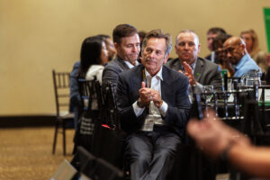 Brampton_Food and Bev Summit_D85_9046 - Invest Brampton | Brampton Economic Development Man in a dark suit clapping at a roundtable event, with other attendees seated and watching in the background