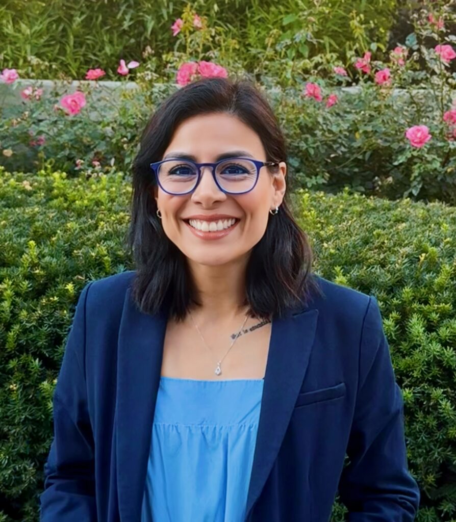 Portrait of a smiling woman with blue-framed glasses wearing a navy blazer, standing in a rose garden.