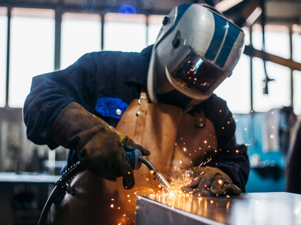 Welder in protective gear and apron sparks flying as they weld metal with a torch in a workshop environment.