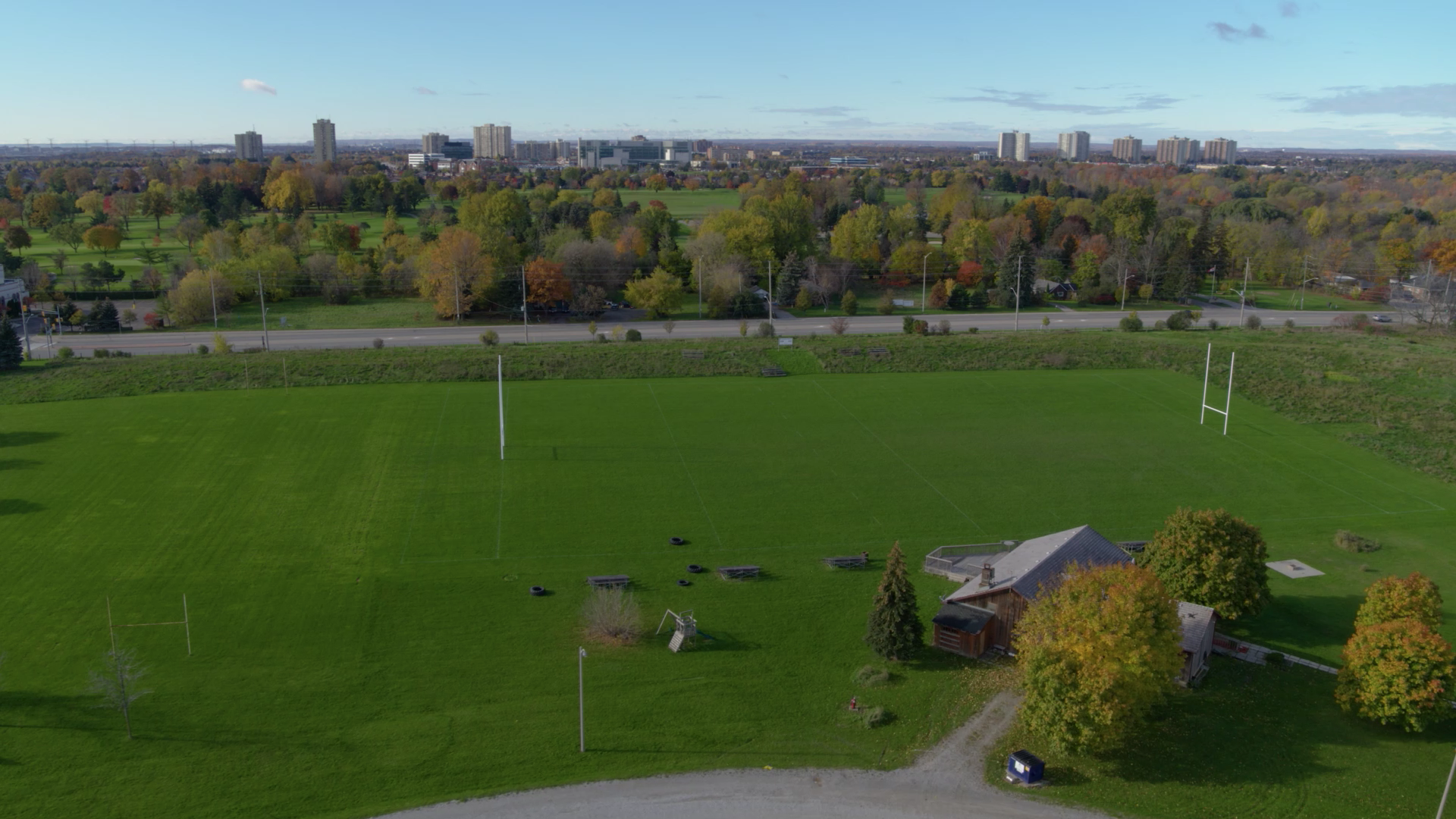 Aerial view of a green rugby field with tall goalposts, nearby trees, and a distant city skyline under a clear blue sky.