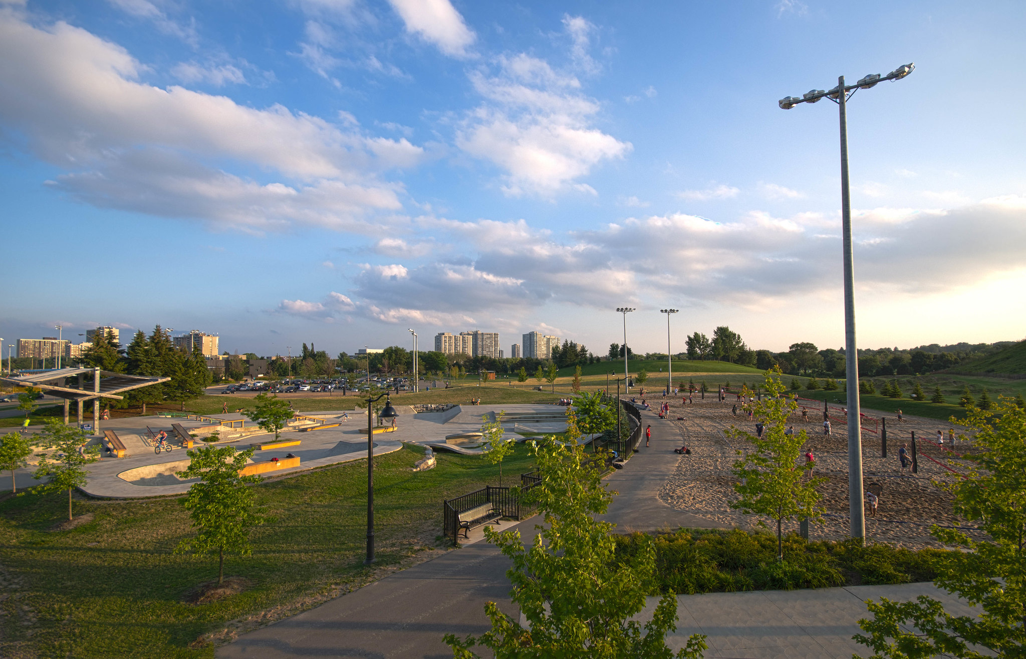 Sunlit urban park with a skate area, winding paths, and a tall floodlight on the right; city buildings in the distance under a blue sky with clouds.