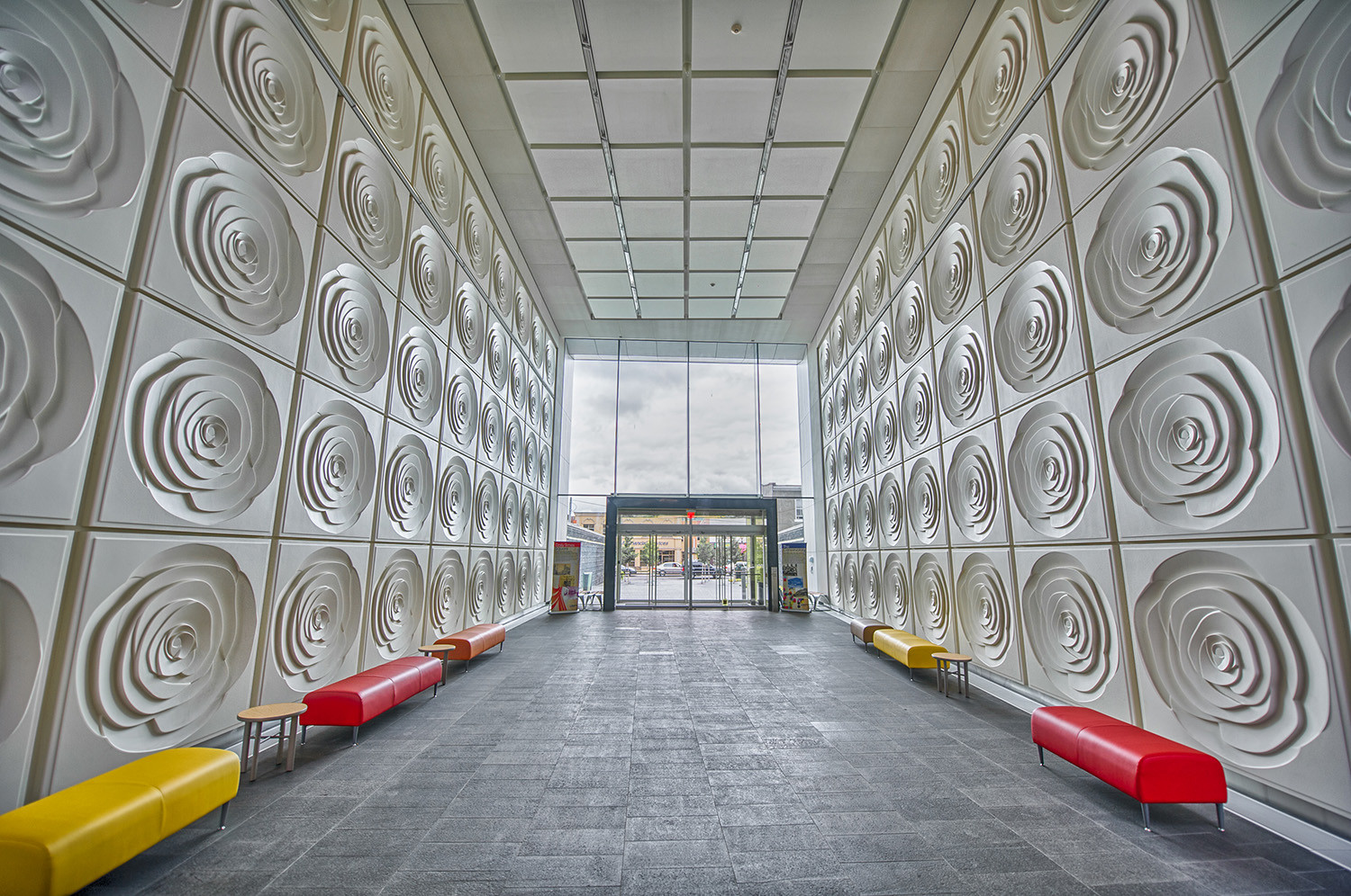 Long modern lobby hallway with white rose-pattern wall panels, gray tiled floor, and colorful red, yellow, and orange benches near the walls.