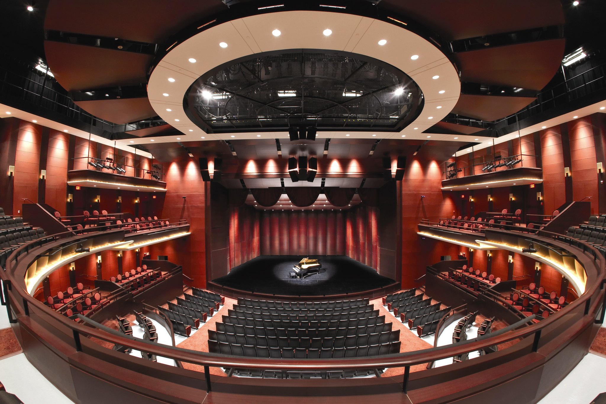Wide view of a modern concert hall with red walls, multiple seating levels, and a black grand piano on a lit stage.