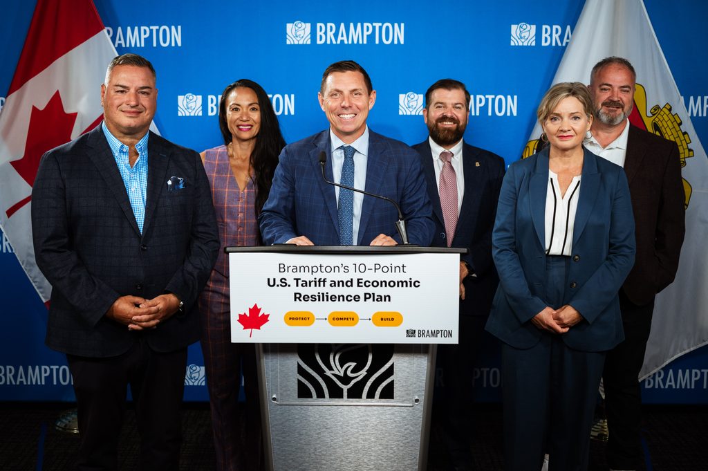 Six professionals in business attire stand behind a podium at a Brampton press conference promoting a 10-point U.S. Tariff and Economic Resilience Plan.