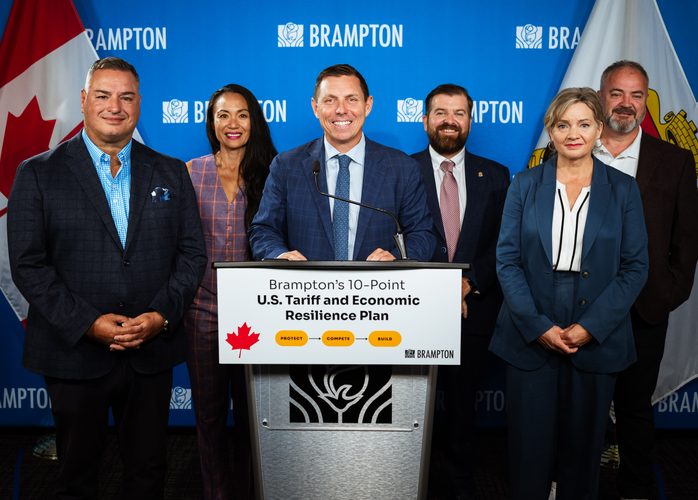 Six professionals in business attire stand behind a podium at a Brampton press conference promoting a 10-point U.S. Tariff and Economic Resilience Plan.
