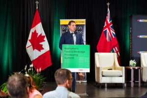 Brampton_Food and Bev Summit_Z62_0012 - Invest Brampton | Brampton Economic Development Male speaker at a podium during a conference, Canadian flag on his left and a Union Jack flag on his right, with banners readout behind him and audience in foreground.