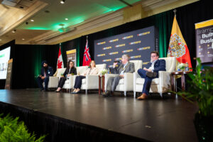 Brampton_Food and Bev Summit_Z62_0184 - Invest Brampton | Brampton Economic Development Panel of five professionals seated on a stage during a conference, branded backdrop reading'Invest Brampton' behind them with flags nearby.