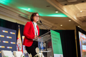 Brampton_Food and Bev Summit_Z62_0272 - Invest Brampton | Brampton Economic Development Woman in a red blazer speaks at a conference podium with microphones and green backstage lighting behind her