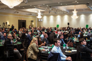 Brampton_Food and Bev Summit_Z62_0367 - Invest Brampton | Brampton Economic Development Crowded conference hall with round tables and many attendees in business attire, green menus on the tables under chandeliers.