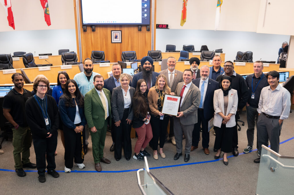Group of diverse professionals posing in a council chamber, holding a framed proclamation while others smile, with flags and a large screen behind.