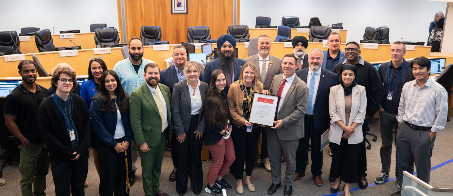 Group of diverse professionals posing in a council chamber, holding a framed proclamation while others smile, with flags and a large screen behind.