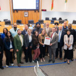Group of diverse professionals posing in a council chamber, holding a framed proclamation while others smile, with flags and a large screen behind.