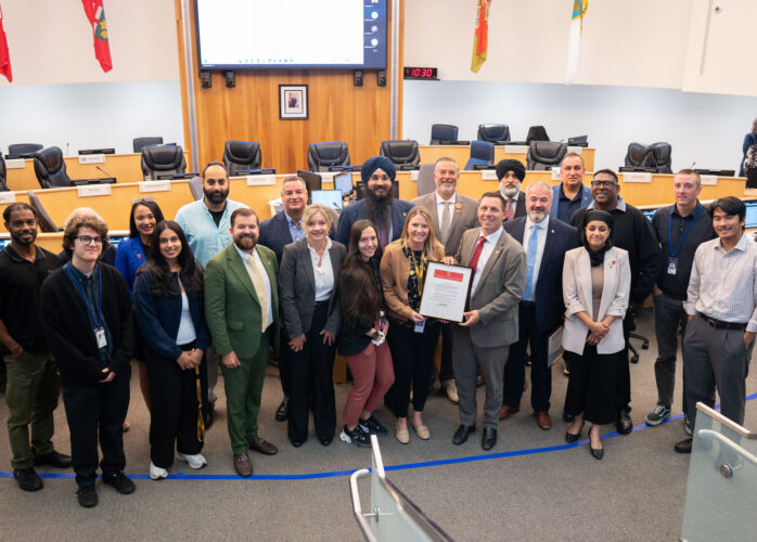 Group of diverse professionals posing in a council chamber, holding a framed proclamation while others smile, with flags and a large screen behind.