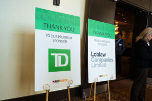 Brampton_Food and Bev Summit_Z62_9698 - Invest Brampton | Brampton Economic Development Two standing event signs with a bright green'THANK YOU' banner and sponsor logos, set up in a conference hall (people in background).