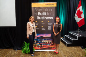 Brampton_Food and Bev Summit_Z62_9860 - Invest Brampton | Brampton Economic Development Two women pose beside a Brampton business banner on a stage, with a Canadian flag nearby.